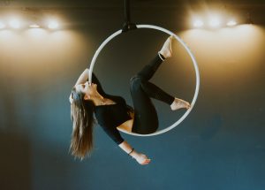 Zajęcia sportowe - Pole Dance i Aerial Hoop. Na zdjęciu ubrana na czarno kobieta z długimi włosami na zawieszonym na linie hula hop.